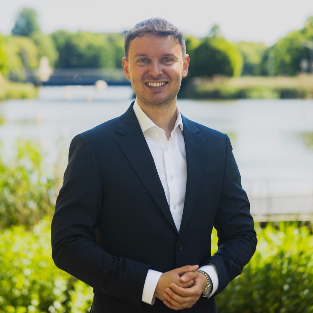 Leo Jürgensen Mann in Anzug lächelt am Ufer eines Sees, umgeben von grüner Landschaft.