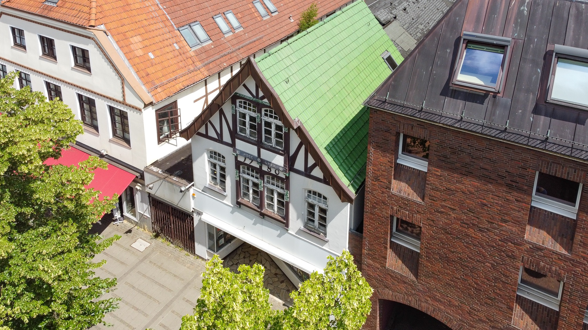 Fachwerkhaus mit grünem Dach neben modernen Gebäuden und gepflastertem Platz.